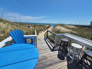 A wooden deck with a blue chair and table, a railing, and a view of the beach and dunes. - SandSpur Ocean Cottages (Nags Head)