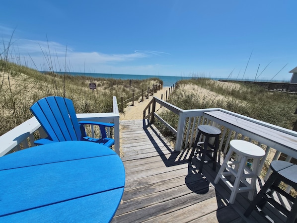 Eine Holzterrasse mit blauem Stuhl und Tisch, einem Geländer und Blick auf Strand und Dünen.