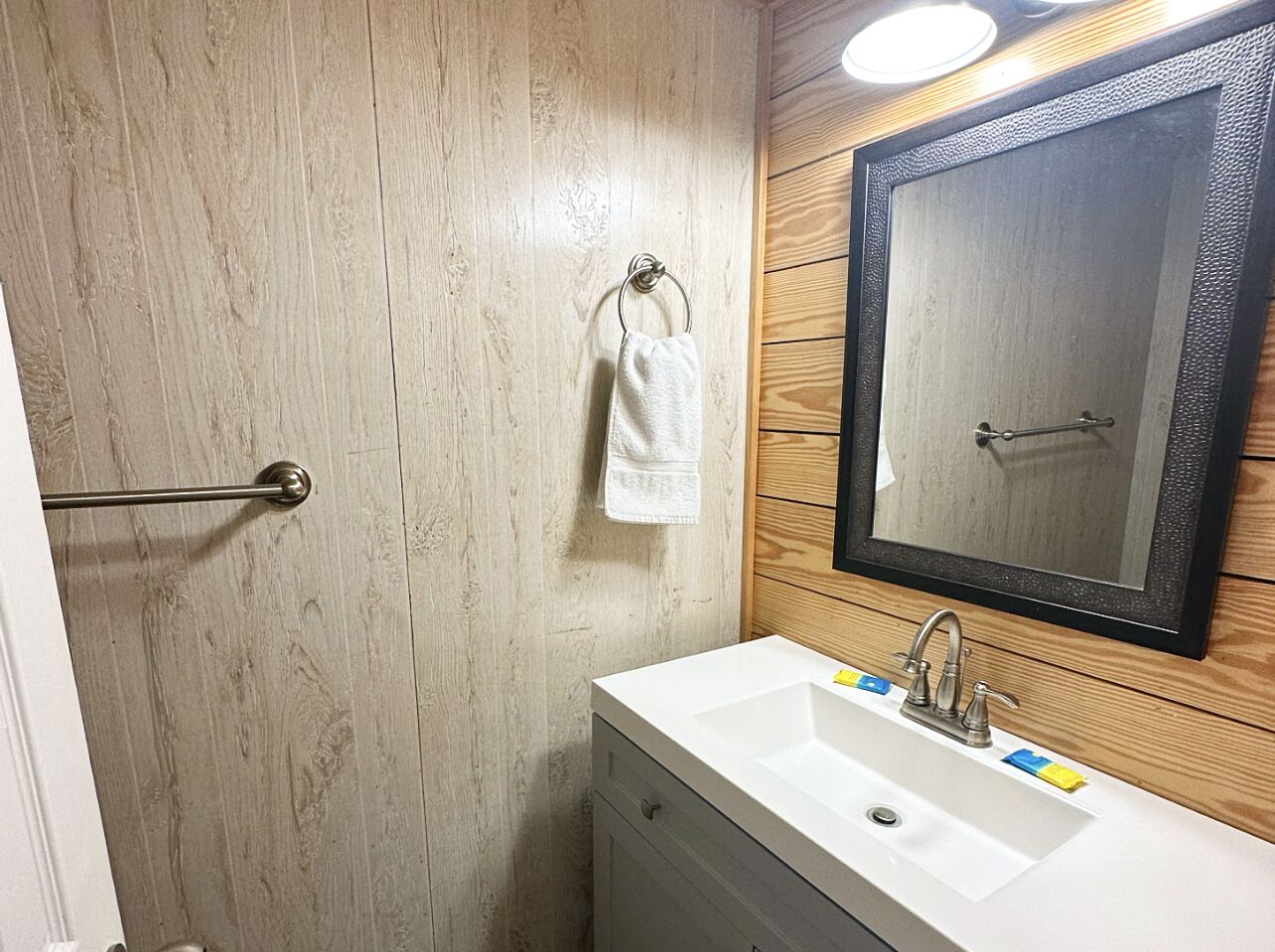 Une salle de bain avec un mur en bois, un lavabo blanc, un miroir et une serviette suspendue à une tringle en métal.