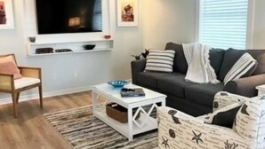 Un saló amb un sofà gris, una taula de centre blanca, un butac armchair estampat, una televisió penjada a la paret i un quadre emmarcat sobre el sofà.