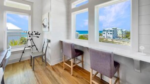 A room with a telescope, two chairs, a table, and large windows with a view of the beach and buildings. - The Barracuda - Cape San Blas (Port Saint Joe)