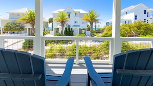 A covered outdoor seating area with two blue chairs, a white pergola, and a view of white buildings with palm trees. - The Barracuda - Cape San Blas (Port Saint Joe)