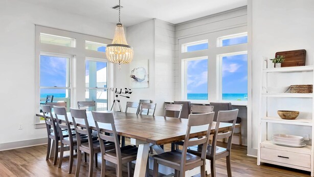 A dining area with a wooden table and chairs, large windows with ocean views, a chandelier, and a white shelving unit with decorative items. - The Barracuda - Cape San Blas (Port Saint Joe)