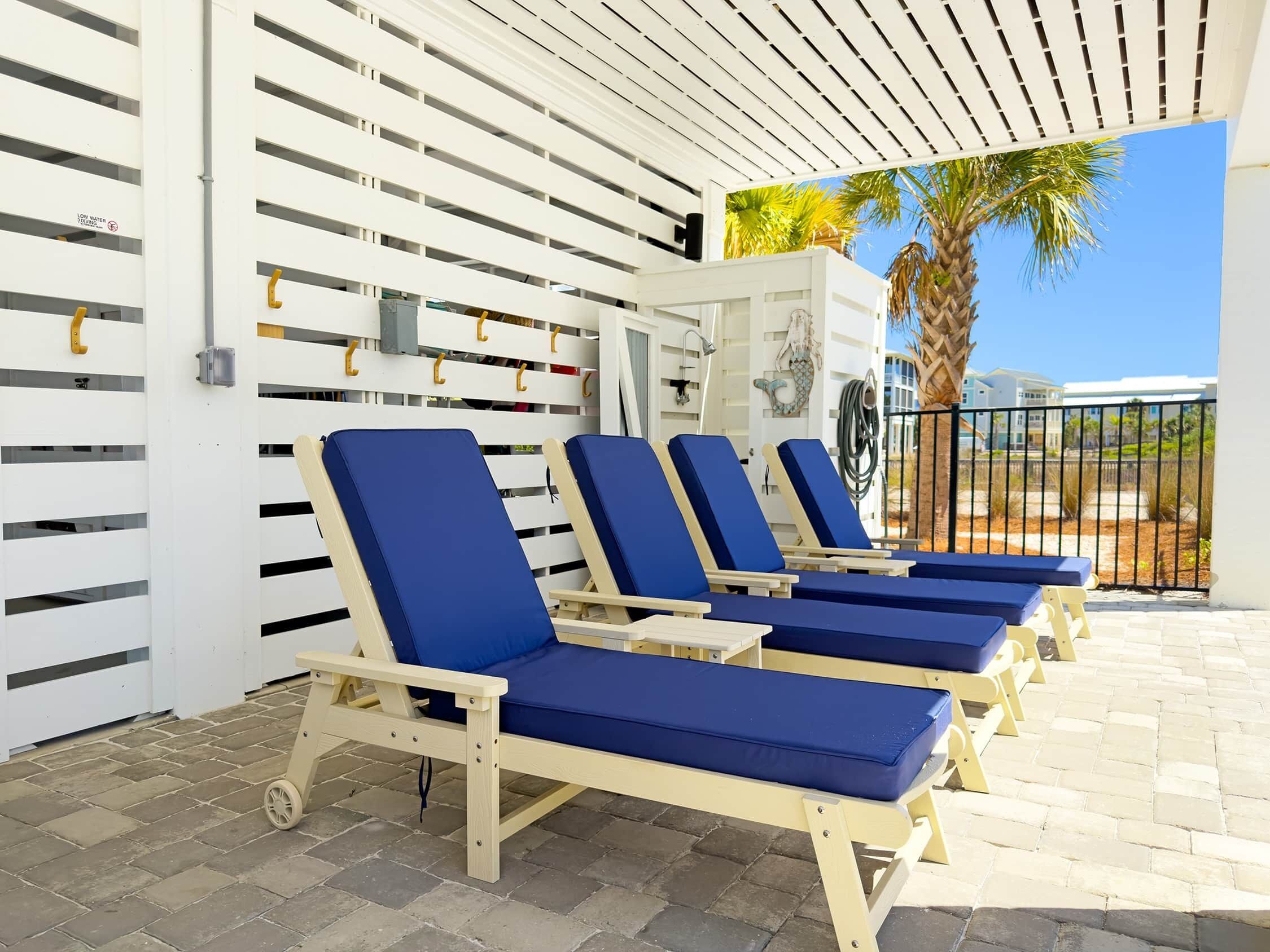 A covered outdoor seating area with lounge chairs, a white wall with horizontal slats, and a clear view of palm trees and buildings in the background.