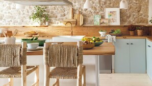 A kitchen with a stone wall, wooden ceiling beams, and a central island with chairs. - Country House North of Spain (Lamadrid)