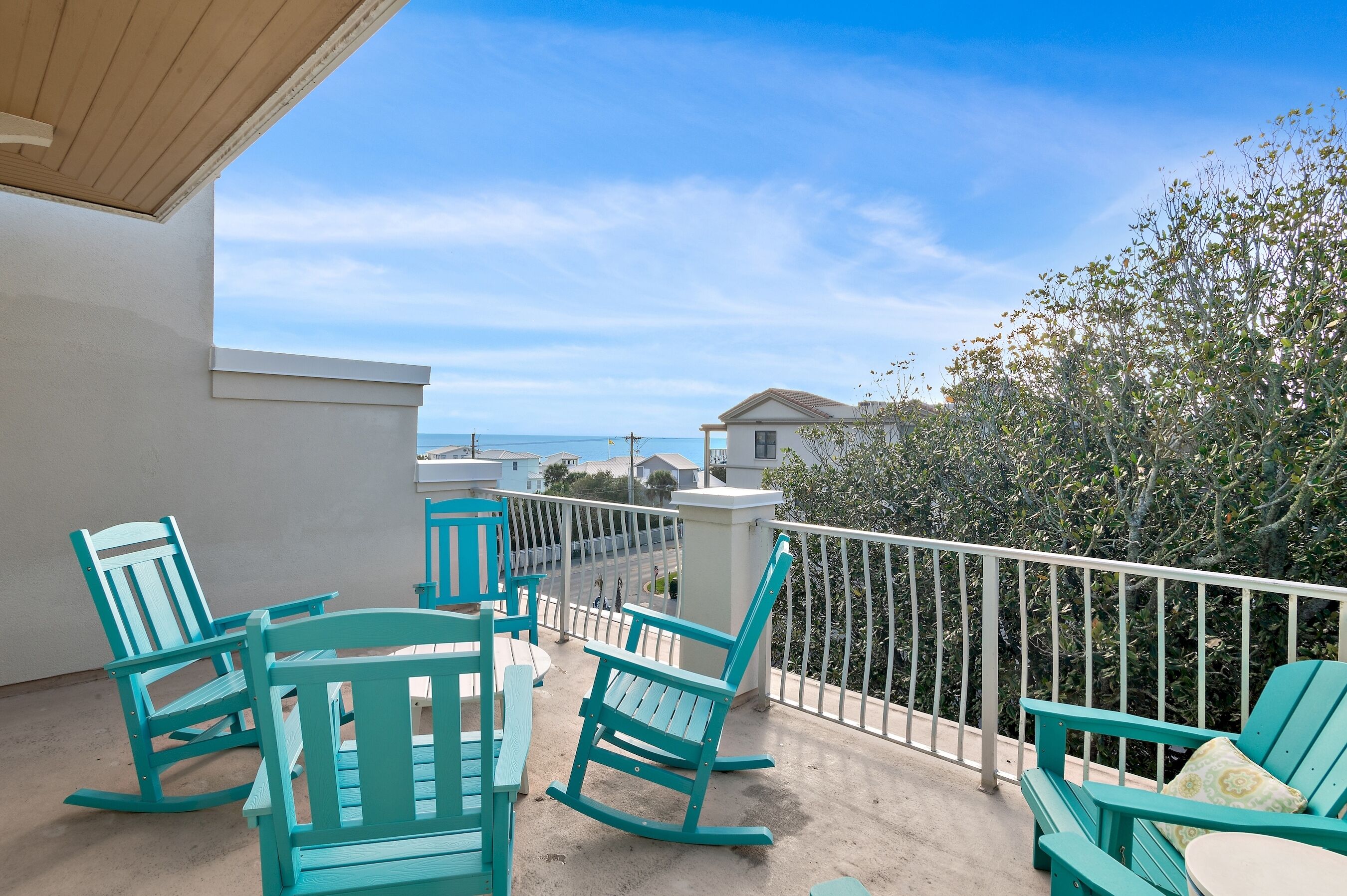 A balcony with turquoise rocking chairs and a table, overlooking a beach and residential buildings.