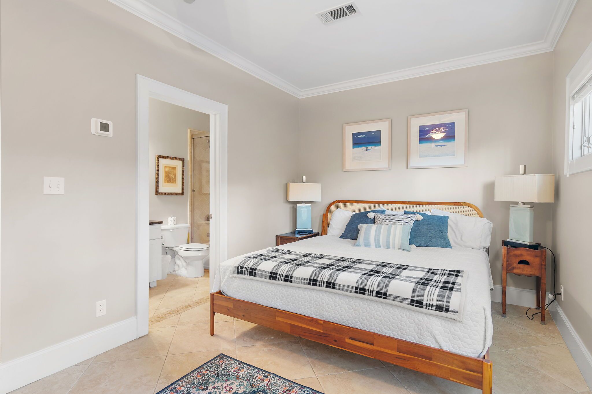 Une chambre à coucher avec un lit en bois, deux tables de chevet avec des lampes, une salle de bain visible à travers une porte ouverte, et deux tableaux encadrés accrochés au mur.