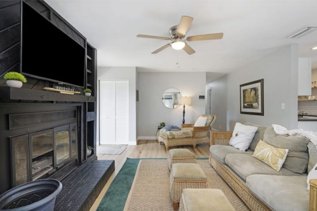 Un salon avec une cheminée, un ventilateur de plafond, un canapé et un pouf en osier.
