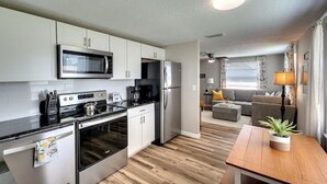 A compact kitchen with modern appliances, a dining area with a wooden table, and a living room with a sofa and a lamp.