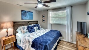 A bedroom with a bed, a ceiling fan, a window with curtains, a nightstand, and a TV on a wooden dresser.