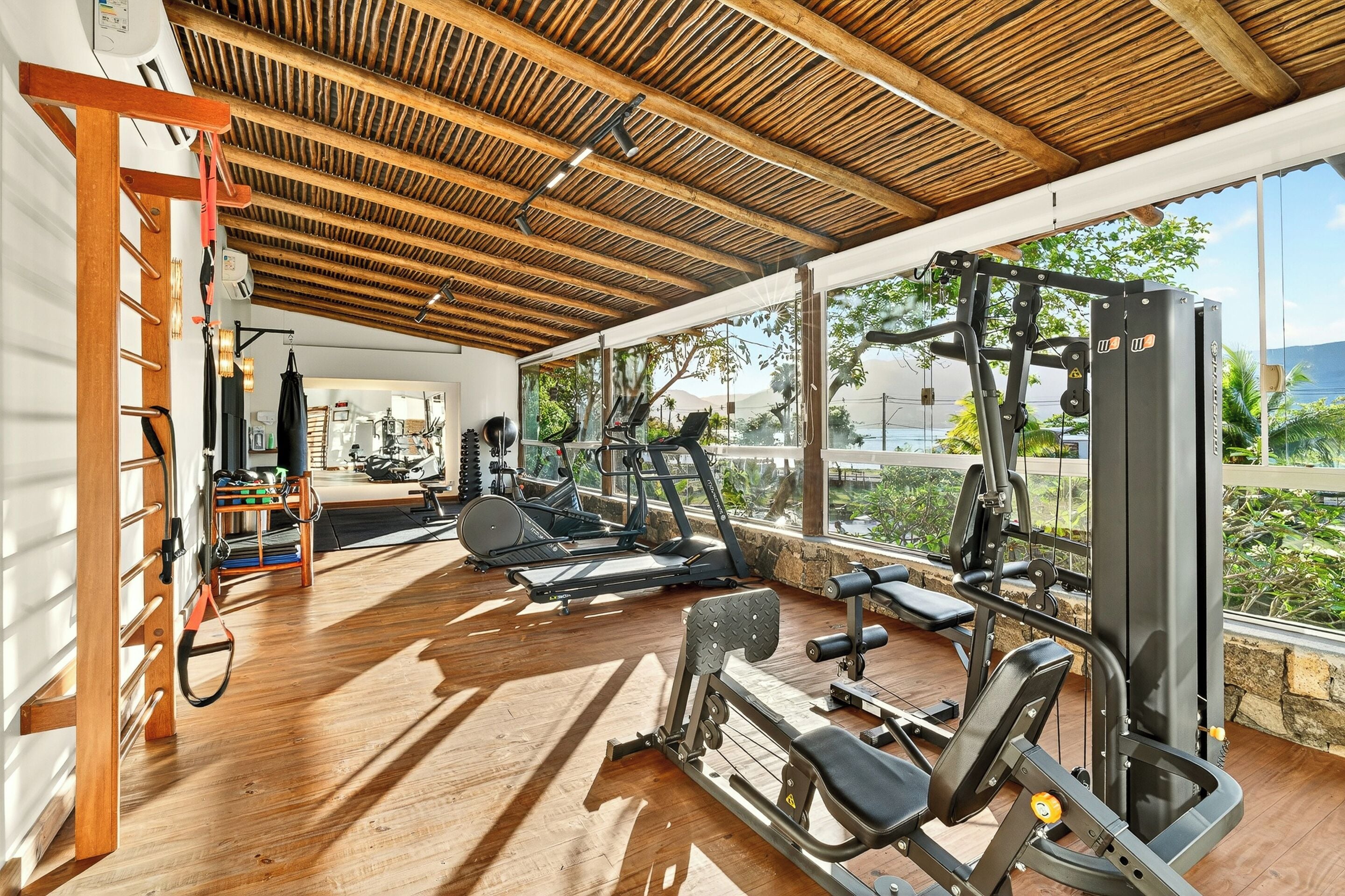 A gym with exercise equipment, a wooden ceiling, and large windows overlooking greenery.