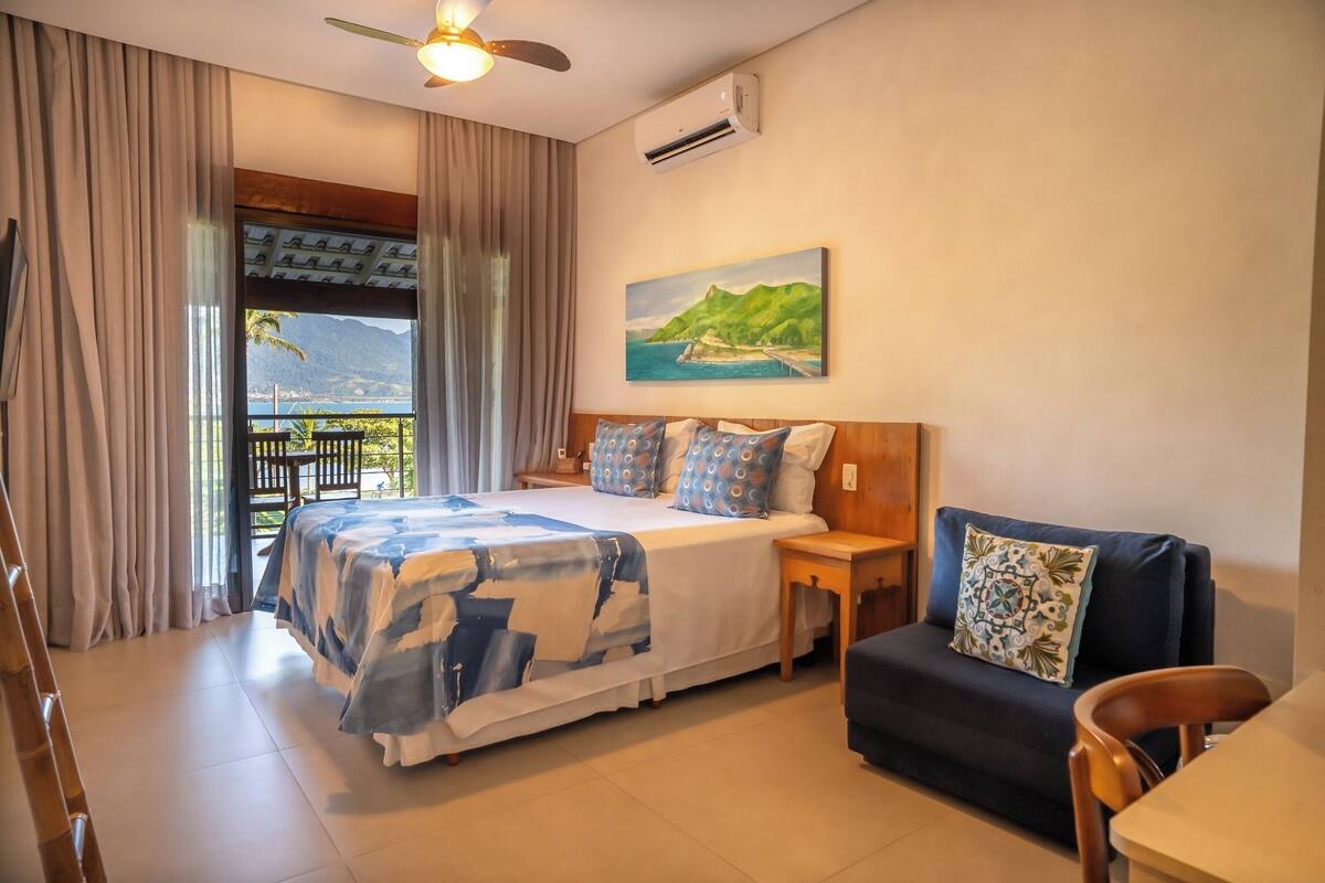 A bedroom with a bed, a sofa, a chair, a painting, and a view of a landscape.