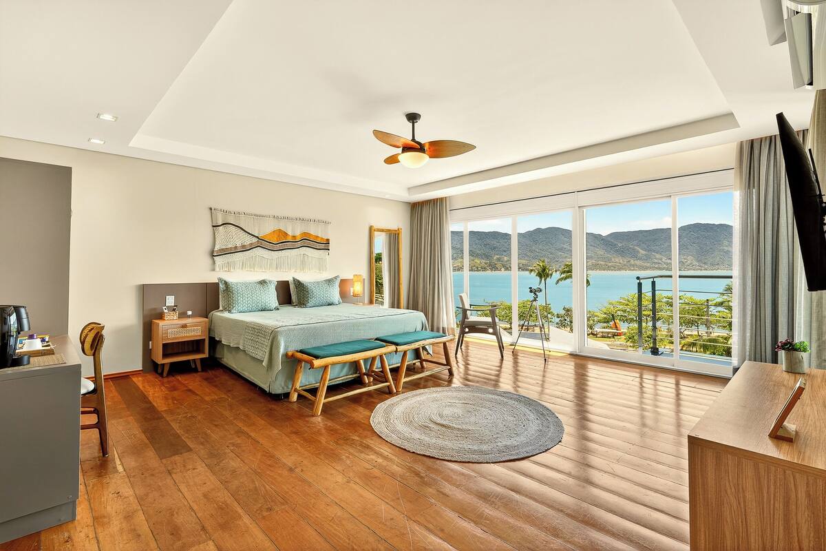 A modern bedroom with a large bed, a desk, and a view of the sea.