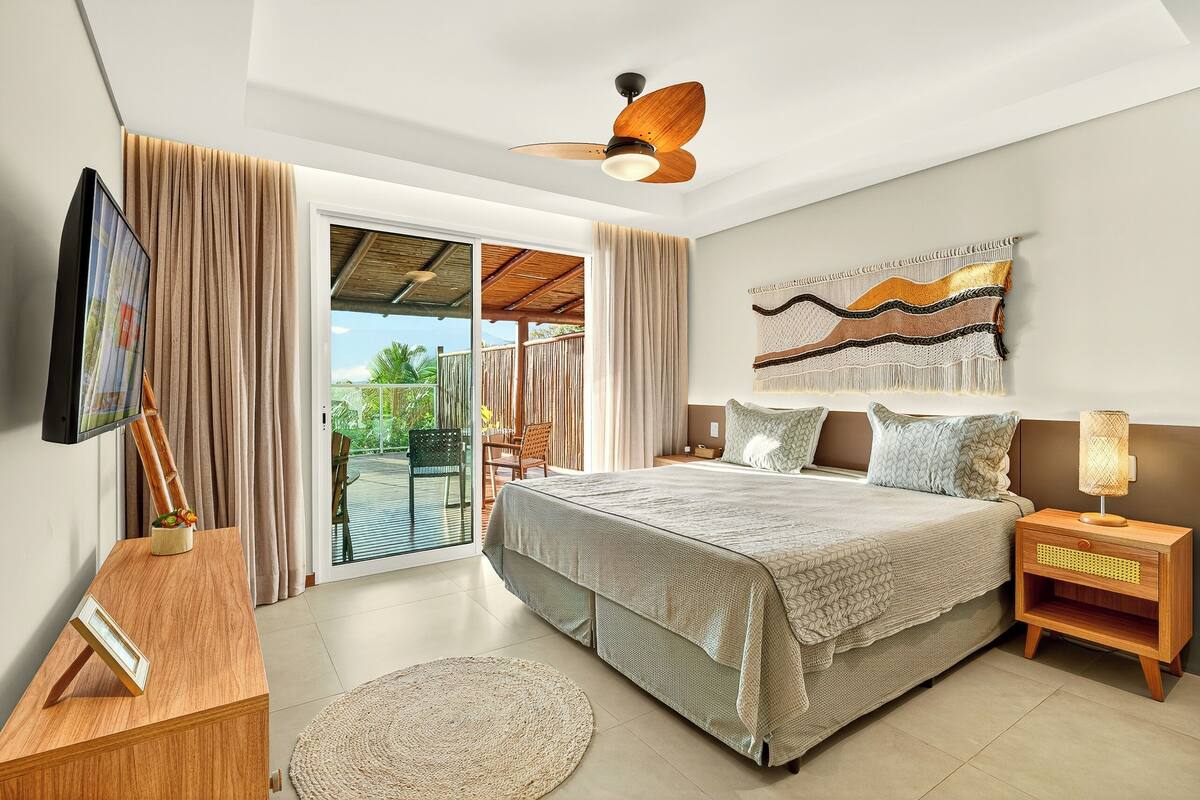 A bedroom with a bed, a TV, a ceiling fan, a nightstand, and a view of the outdoors.