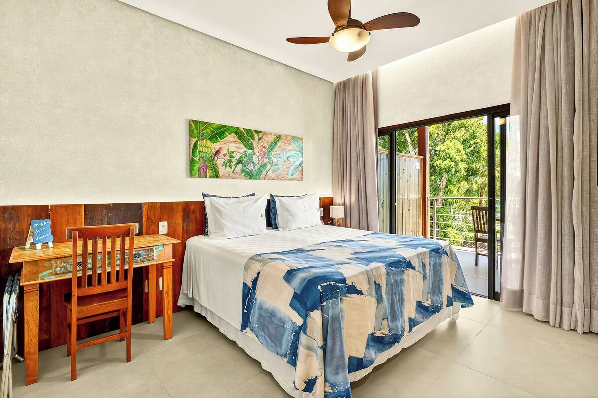 A bedroom with a bed, a desk, and a chair. There is a painting on the wall and a window with curtains.