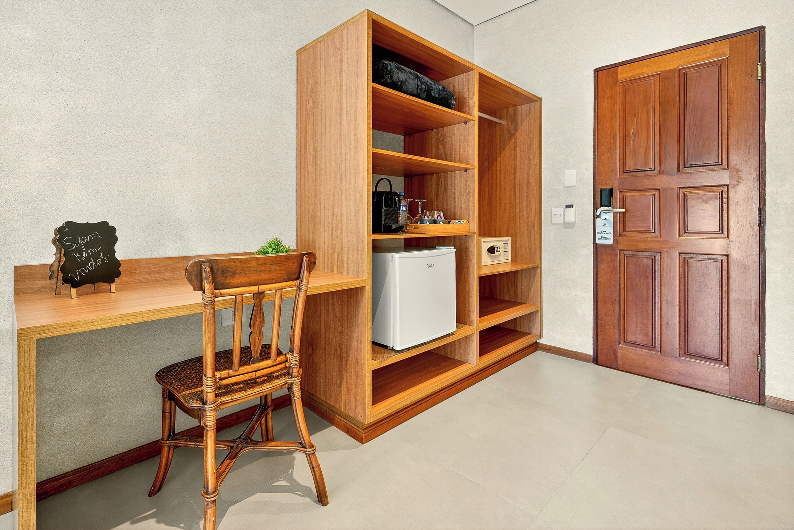 A room with a wooden desk, a wicker chair, a white refrigerator, and a wooden wardrobe.
