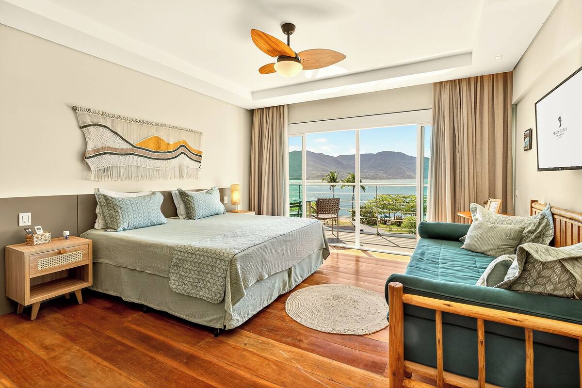 A bedroom with a bed, a sofa, a nightstand, a ceiling fan, a large window with a view of the sea and mountains, and a wall-mounted artwork.