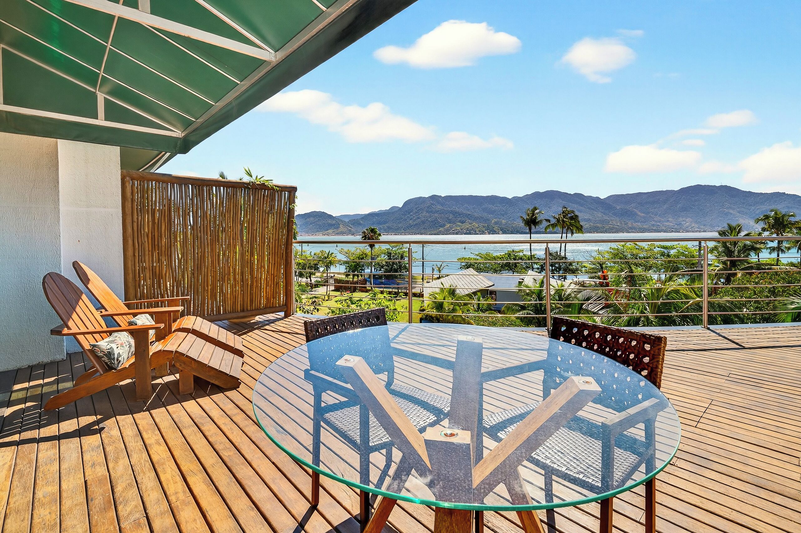 Una terraza de madera con una mesa y sillas de vidrio, con vistas a un cuerpo de agua y montañas.