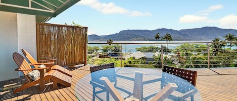Una terraza de madera con una mesa y sillas de vidrio, con vistas a un cuerpo de agua y montañas.