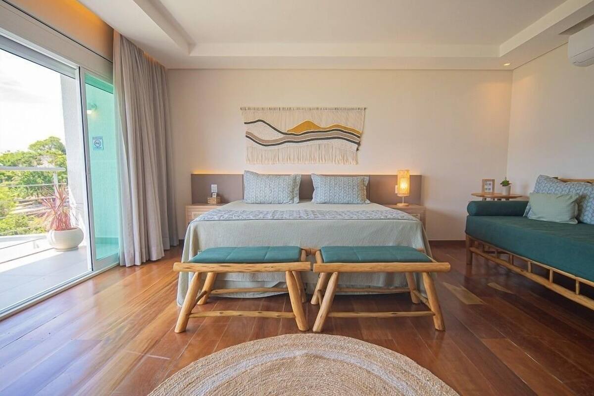 A modern bedroom with a large bed, a sofa, a wooden bench, and a view of greenery through the window.