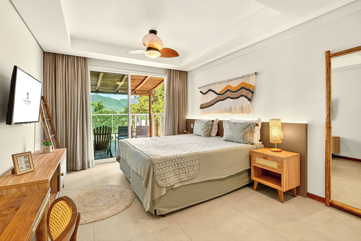 A bedroom with a bed, bedside table, lamp, and a large mirror. There is a window with curtains, a ceiling fan, and a wall-mounted artwork.