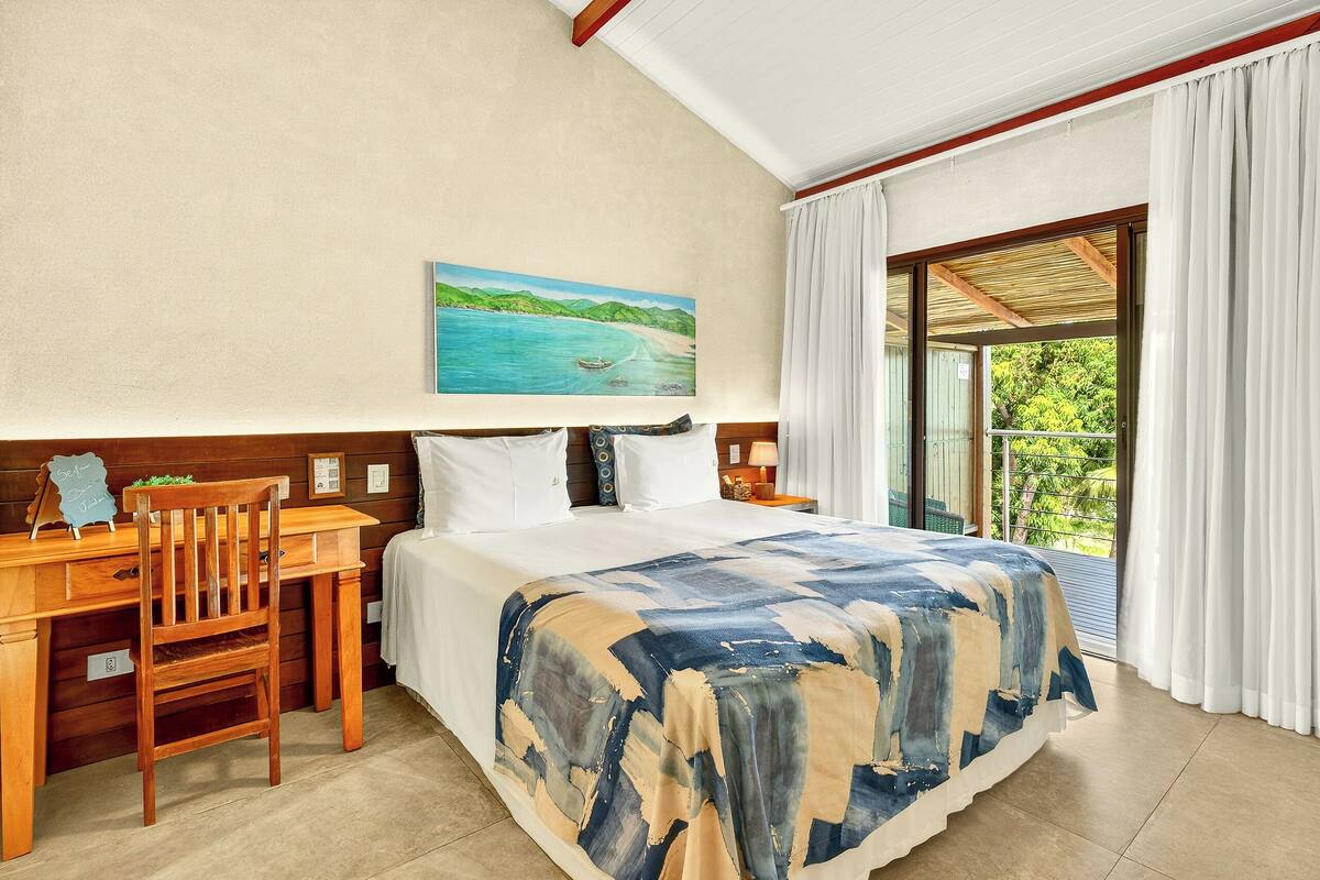 A hotel room with a bed, a desk, a chair, a painting of a beach, and a balcony with curtains.