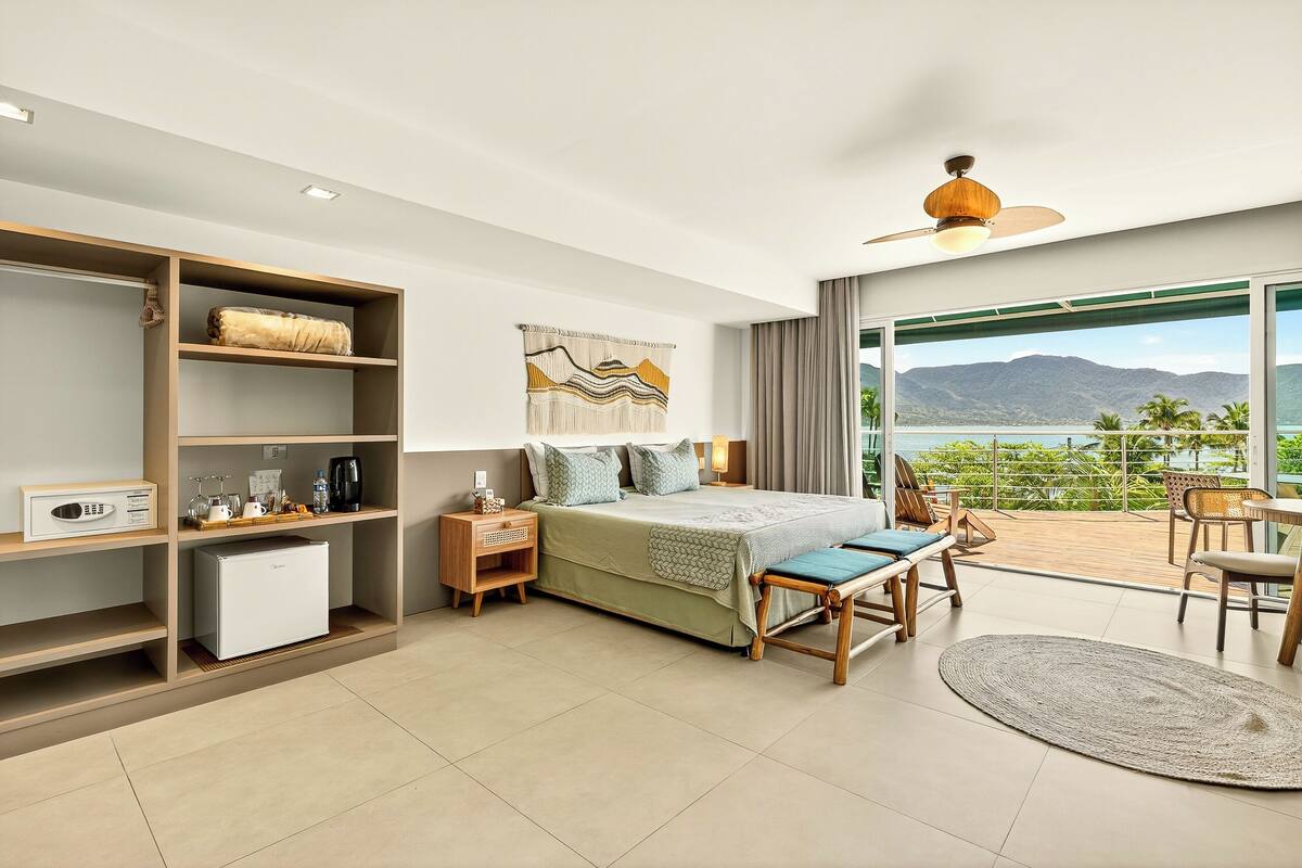 A modern hotel room with a bed, a small kitchenette, a balcony with a view, and a ceiling fan.