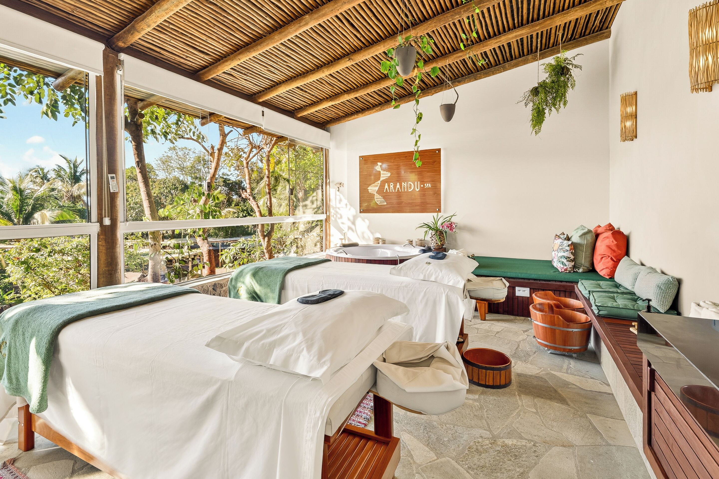A spa room with two beds, a massage table, a bench, and a view of the outdoors.