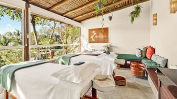 A spa room with two beds, a massage table, a bench, and a view of the outdoors.