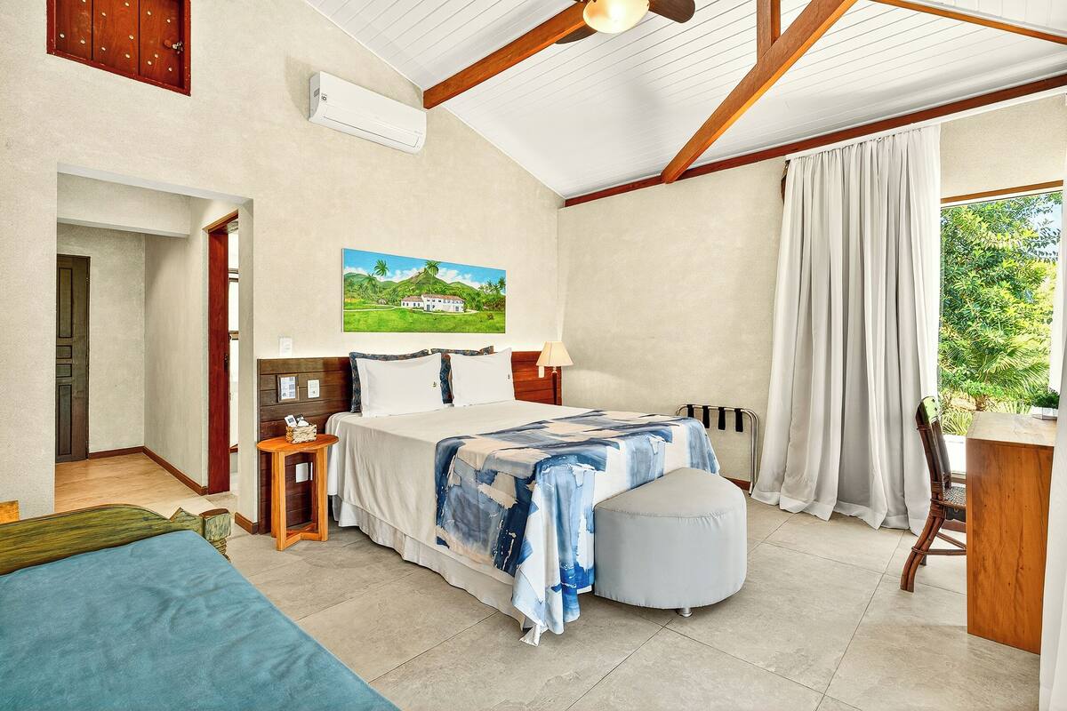 A bedroom with a bed, a desk, a chair, a small table, and a window with curtains.