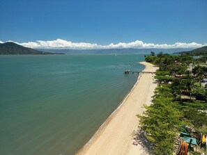 A beach with a pier extending into calm waters, surrounded by greenery and buildings.