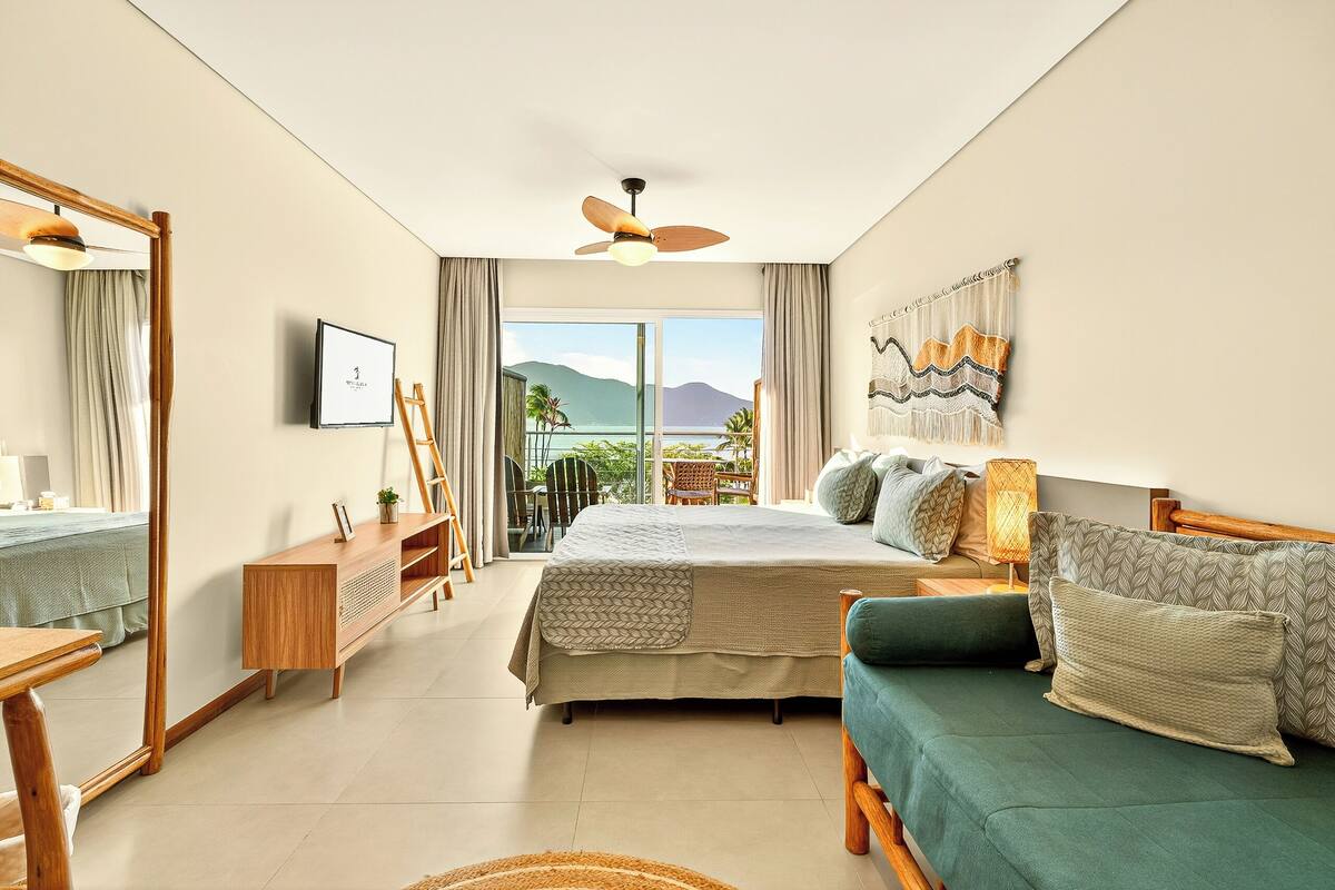 A bedroom with a bed, a sofa, a wooden dresser, a TV, and a large window with a view of mountains and the sea.