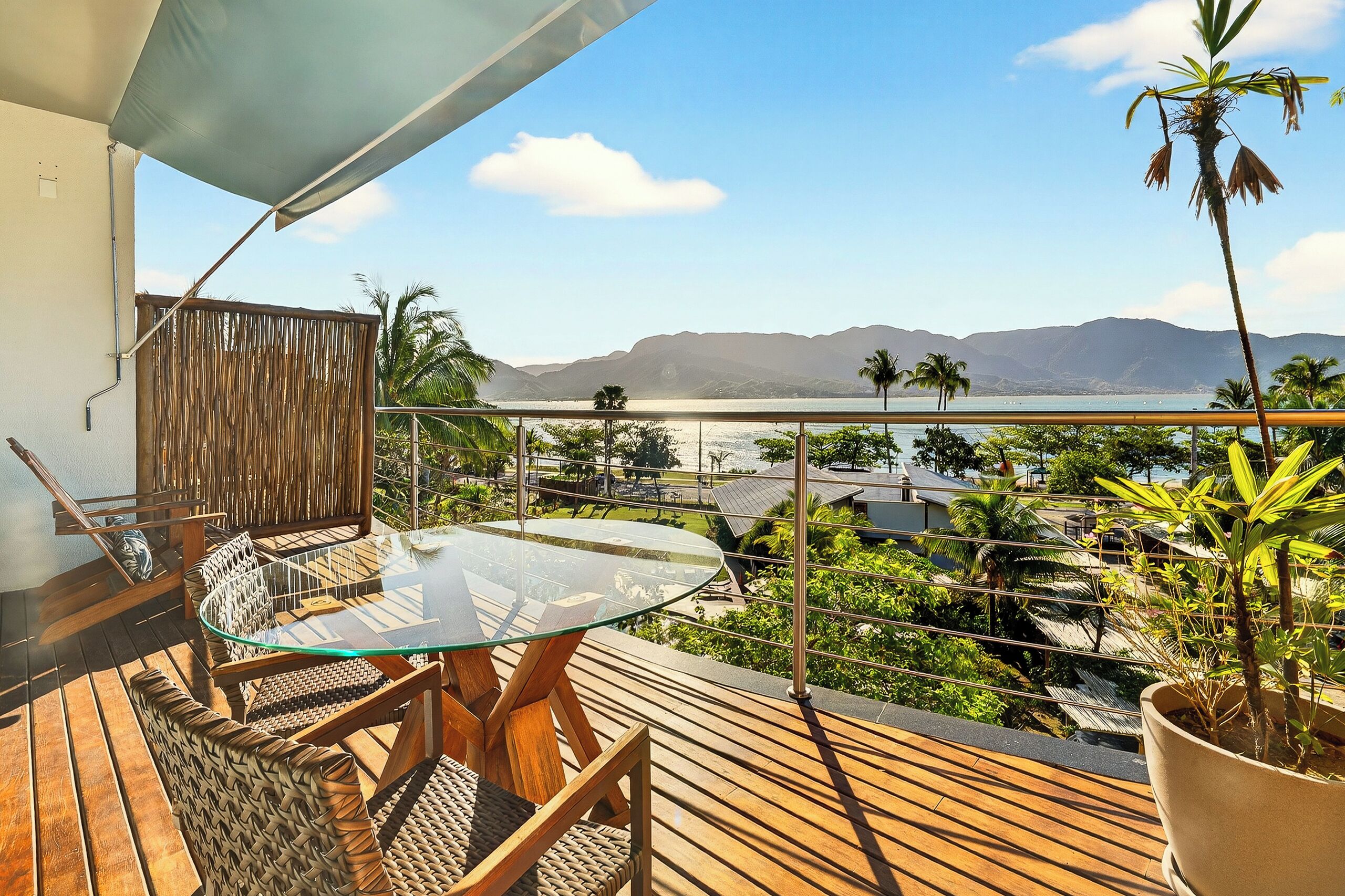 Un balcon doté d’une table en verre et de chaises en osier, offrant une vue panoramique sur les montagnes et un plan d’eau.