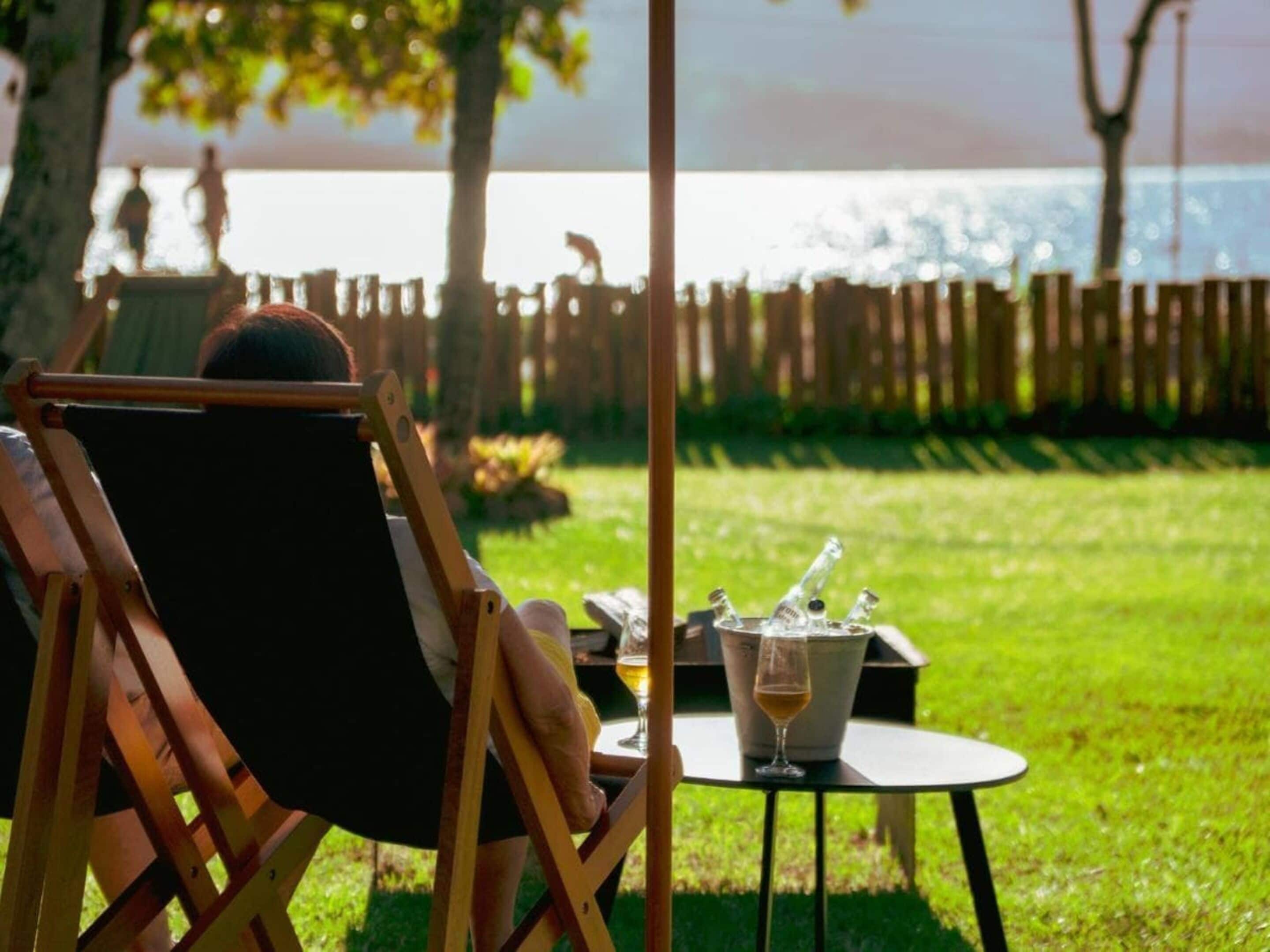 A person relaxing in a lawn chair with a view of the sea, a table with a drink, and a bucket of ice.