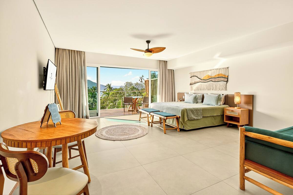 A modern hotel room with a bed, a dining table, chairs, a bench, a ceiling fan, and a view of the outdoors.