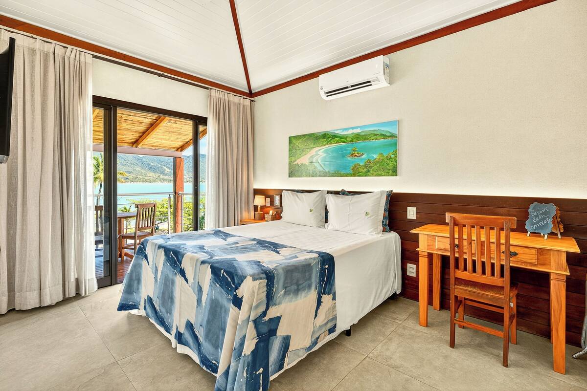 A bedroom with a bed, a desk, a chair, a painting, and a view of the outdoors.