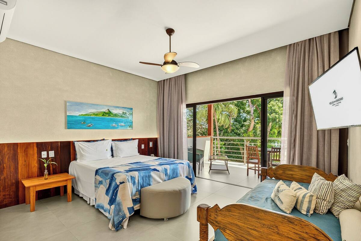 A bedroom with a bed, a wooden bench, a ceiling fan, a painting, and a balcony with a chair.