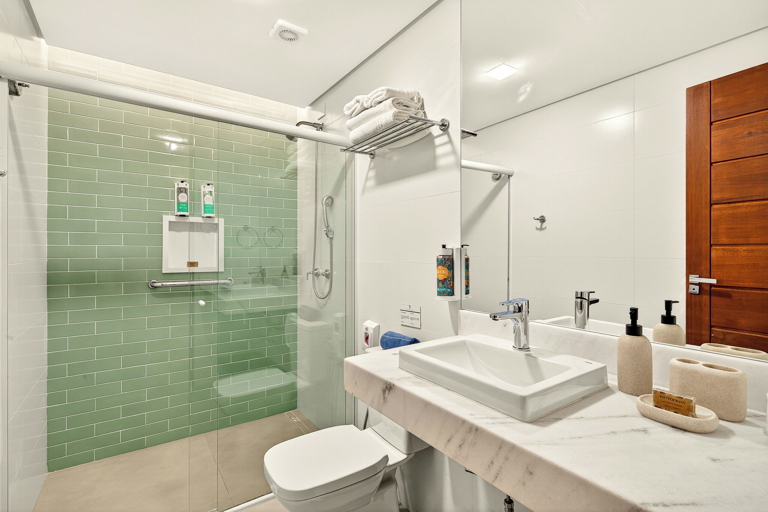 A modern bathroom with a glass shower enclosure, a white toilet, a marble sink with toiletries, and a wooden door.