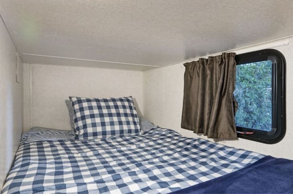 Pequeña habitación con cama individual, colcha a cuadros, ventana con cortinas y vistas a zonas verdes.