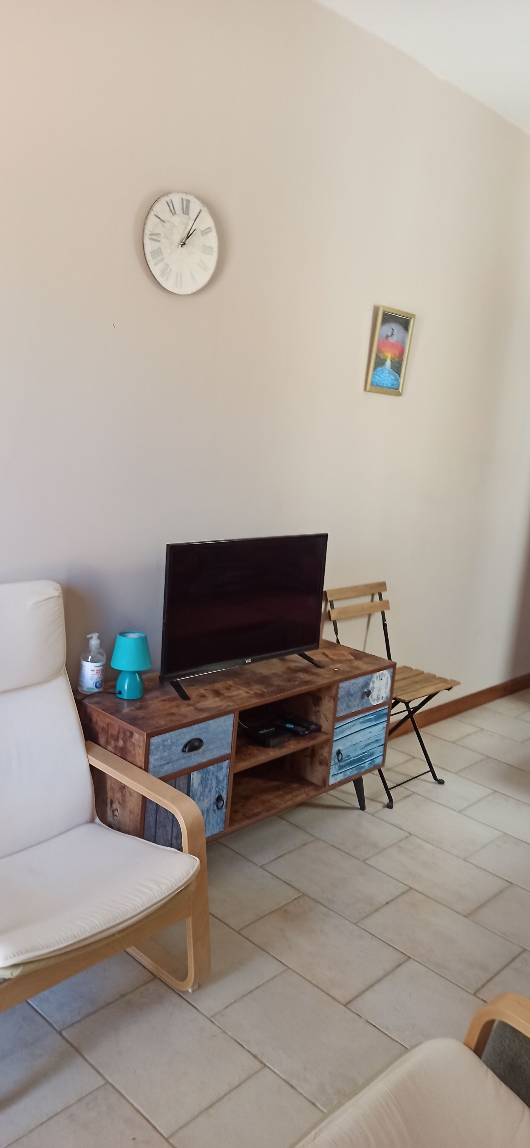 Een woonkamer met een houten tv-meubel, een witte stoel, een klok en een klein, ingelijst foto op de muur.