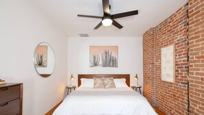 A bedroom with a wooden floor, a bed with white bedding, a ceiling fan, and a brick wall. - Downstream Loft - Historic Savory Building (Salida)
