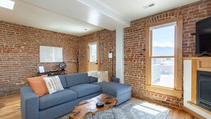A living room with a brick wall, a blue sectional sofa, a wooden coffee table, and a fireplace. - Downstream Loft - Historic Savory Building (Salida)