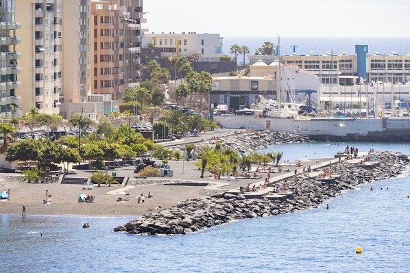 Zona costera con un rompeolas rocoso, playa y edificios al fondo.