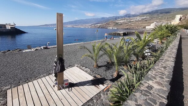 Ein Strand mit einer Holzterrasse, felsigem Ufer und Blick auf das Meer und ferne Hügel.