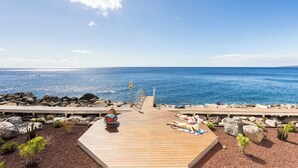 Eine Holzterrasse mit zwei Liegestühlen und einem kleinen Tisch, mit Blick auf eine felsige Küste und das Meer.