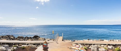A wooden deck with two sun loungers and a small table, overlooking a rocky shoreline and the sea.
