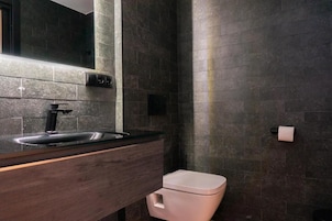 Un baño moderno con pared de azulejos oscuros, tocador de madera, lavamanos y retrete.
