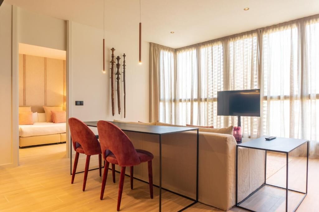 Una sala de estar moderna con un sofá, una mesa de comedor y dos sillas rojas. Hay un televisor sobre un mueble bajo y una lámpara sobre la mesa.