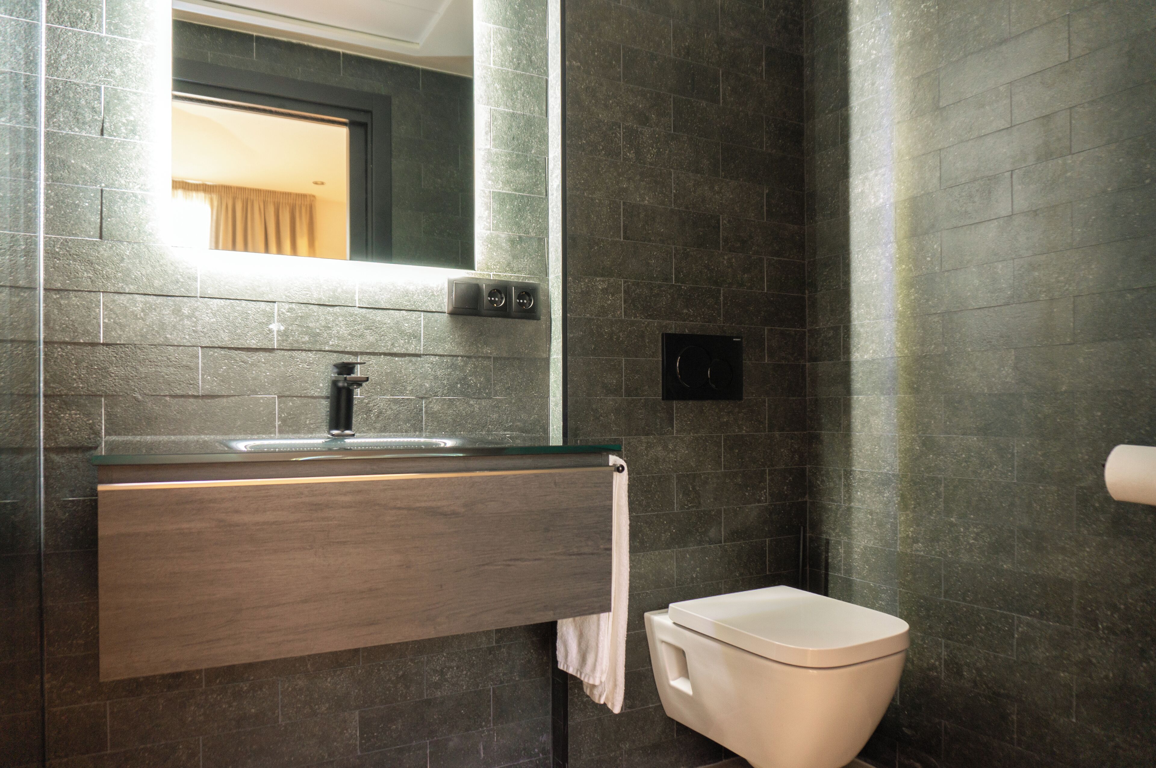 A modern bathroom with a dark tiled wall, a wooden vanity with a sink, a mirror, and a toilet.