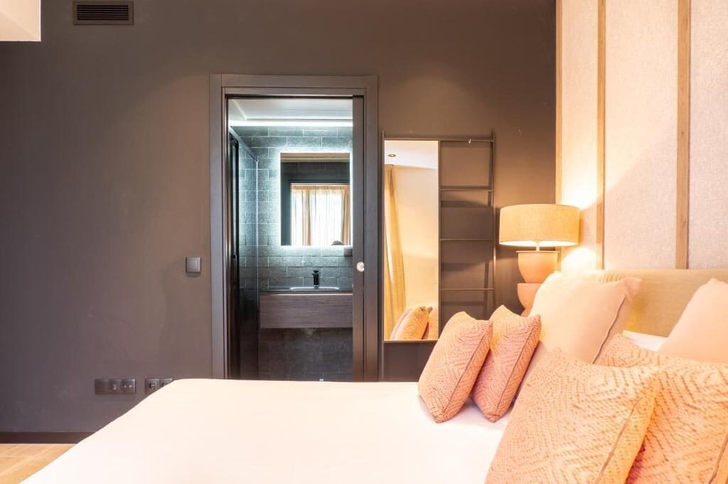 Habitación de hotel con cama, lámpara de noche, espejo y vistas al baño.